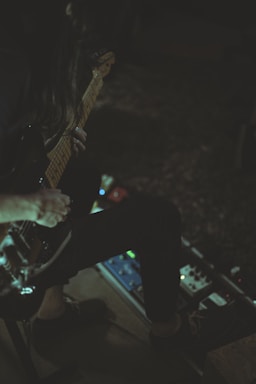 A person is seated, playing an electric guitar with one hand on the neck of the instrument. Various guitar pedals are spread out on the floor near their feet, suggesting the creation of music or sound experimentation. The lighting is dim, casting shadows and giving the scene an intimate atmosphere.