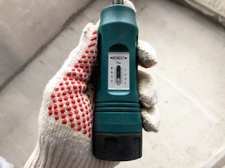 A hand wearing a white glove with red dotted patterns is holding a green torque wrench. The wrench has a black grip and a white dial displaying numerical settings ranging from 1 to 8 marked as 'MOMENT'. The background is a blurred concrete surface.