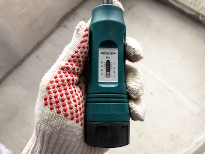 A hand wearing a white glove with red dotted patterns is holding a green torque wrench. The wrench has a black grip and a white dial displaying numerical settings ranging from 1 to 8 marked as 'MOMENT'. The background is a blurred concrete surface.