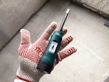 A hand wearing a red and white dotted work glove is holding a green moisture meter. The device, featuring a small display, is against a concrete background.