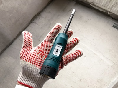A hand wearing a red and white dotted work glove is holding a green moisture meter. The device, featuring a small display, is against a concrete background.