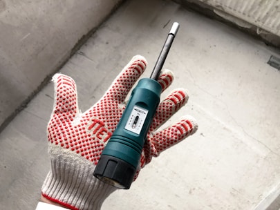 A hand wearing a red and white dotted work glove is holding a green moisture meter. The device, featuring a small display, is against a concrete background.