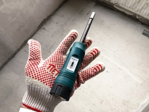 A hand wearing a red and white dotted work glove is holding a green moisture meter. The device, featuring a small display, is against a concrete background.