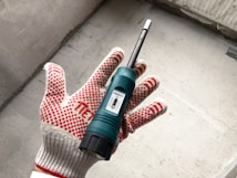 A hand wearing a red and white dotted work glove is holding a green moisture meter. The device, featuring a small display, is against a concrete background.