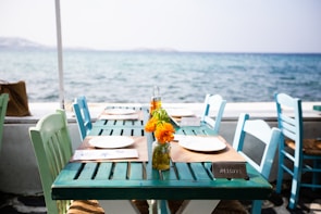 A seaside restaurant setting with a turquoise wooden table and several mismatched pastel chairs. The table is set with white plates on brown placemats, and small glass jars containing vibrant orange flowers serve as centerpieces. In the background, the calm sea stretches out under a clear sky.