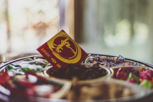 A tray containing various candies and dried fruits is centered, with a red and yellow card featuring calligraphy prominently placed on top. The background is softly blurred, creating a cozy and inviting atmosphere.