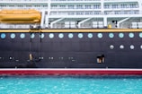 A large cruise ship is docked with a worker in a yellow raincoat standing on a suspended platform, engaged in maintenance or cleaning. The ship features multiple levels with balconies, circular windows, and a lifeboat positioned above. The water below is a striking turquoise color.