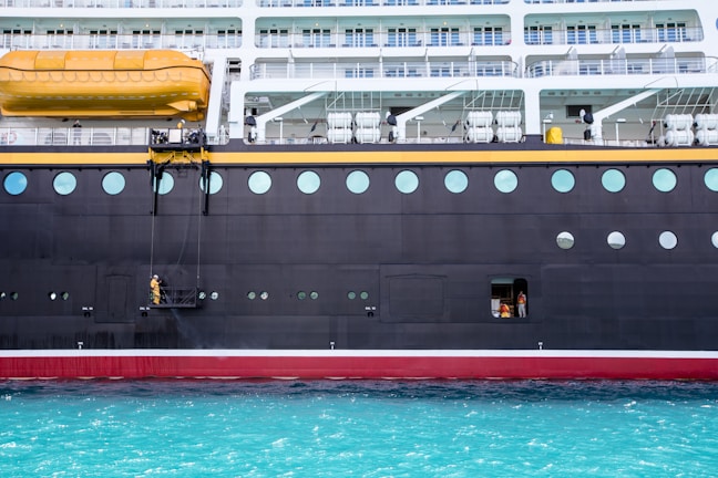 A large cruise ship is docked with a worker in a yellow raincoat standing on a suspended platform, engaged in maintenance or cleaning. The ship features multiple levels with balconies, circular windows, and a lifeboat positioned above. The water below is a striking turquoise color.