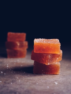 Three stacked orange translucent gummy candies with a sugar coating are placed on a dark surface. The background is dimly lit, accentuating the texture and color of the candies.