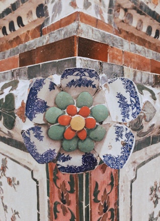 Ornate ceramic flower decoration with blue and white intricate patterns around green and orange circular elements, set against a richly textured architecture background with floral motifs.