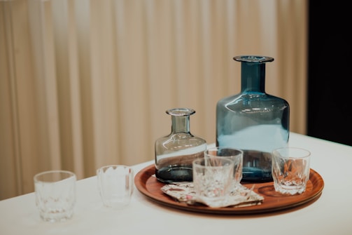 On a table, a wooden tray holds a large blue glass bottle and a smaller gray bottle, surrounded by four clear glass cups. The bottles and cups have simple, elegant designs, and the arrangement looks neat. A patterned cloth is placed on the tray, and the background features light beige curtains.