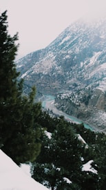 A scenic mountainous landscape with snow-covered peaks and lush green trees. A winding turquoise river flows through the valley, surrounded by rocky cliffs and forested slopes.
