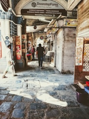 A narrow market alley with stone paving, lined with closed shop shutters and decorated with hanging vinyl records. Signs offering currency exchange and travel services are prominently displayed. Several people are walking through the alley, passing by stalls displaying colorful artwork.