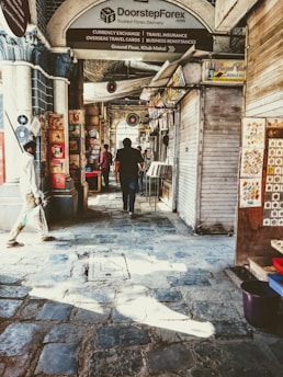 A narrow market alley with stone paving, lined with closed shop shutters and decorated with hanging vinyl records. Signs offering currency exchange and travel services are prominently displayed. Several people are walking through the alley, passing by stalls displaying colorful artwork.