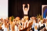 A large crowd is raising their hands in what appears to be an enthusiastic response. In the background, there is a person with their arms raised, possibly a speaker at an event. The scene suggests a lively and interactive atmosphere, indicative of a conference, seminar, or performance.