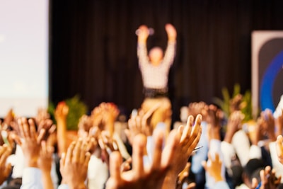 A large crowd is raising their hands in what appears to be an enthusiastic response. In the background, there is a person with their arms raised, possibly a speaker at an event. The scene suggests a lively and interactive atmosphere, indicative of a conference, seminar, or performance.