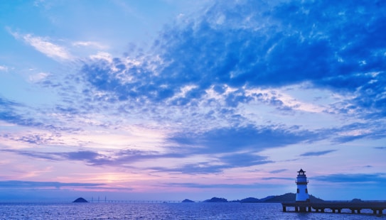 A serene landscape with a lighthouse standing on a pier extending into the water. The sky is expansive, filled with soft, fluffy clouds tinted in hues of blue, pink, and purple as the sun sets or rises. Gentle waves ripple across the ocean, providing a tranquil foreground.