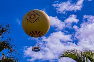 A large yellow hot air balloon decorated with a sun emblem and patterns ascends into a vibrant blue sky filled with scattered white clouds. The lower portion of the balloon, including the passenger basket, is visible. Palm tree leaves are framing the image from the corners.