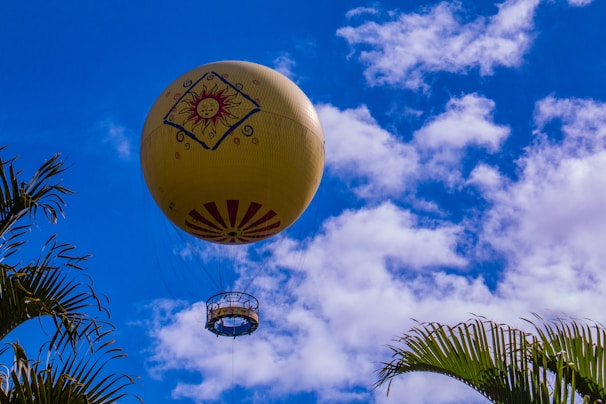 A large yellow hot air balloon decorated with a sun emblem and patterns ascends into a vibrant blue sky filled with scattered white clouds. The lower portion of the balloon, including the passenger basket, is visible. Palm tree leaves are framing the image from the corners.