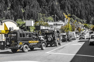 A busy street scene featuring a convoy of safari vehicles parked along the road lined with various shops and businesses. The background is dominated by lush green forested hills. The road is bustling with different types of vehicles, and pedestrians can be seen crossing the street. The scene is lively and vibrant with a mix of natural and urban elements.