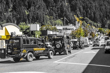 A busy street scene featuring a convoy of safari vehicles parked along the road lined with various shops and businesses. The background is dominated by lush green forested hills. The road is bustling with different types of vehicles, and pedestrians can be seen crossing the street. The scene is lively and vibrant with a mix of natural and urban elements.