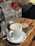 A white cup filled with coffee sits on a saucer placed on a wooden surface. A single red rose in a glass vase adds a touch of elegance beside the coffee cup. The background features a blurred pattern and a candle adds a warm, cozy ambiance.