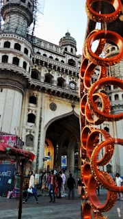 A historic architectural monument with intricate designs and multiple arches is prominently featured. In the foreground, there are vibrant orange bangles displayed on a stand, creating a contrast with the grand structure. People are walking around the area, suggesting a busy market environment.