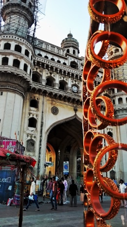 A historic architectural monument with intricate designs and multiple arches is prominently featured. In the foreground, there are vibrant orange bangles displayed on a stand, creating a contrast with the grand structure. People are walking around the area, suggesting a busy market environment.
