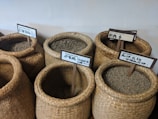Several woven baskets filled with raw coffee beans are arranged closely together. Each basket has a sign with text in multiple languages, indicating different types or origins of the coffee beans. The scene is set against a plain background.