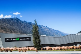 A modern supermarket building with a sign reading 'Countdown Supermarket' is situated at the base of striking, rugged mountains. The architecture of the building is sleek and gray, blending with the natural surroundings. The foreground features decorative grass and a tall tree, set against a clear blue sky.