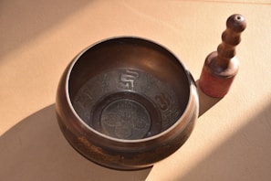 A metal singing bowl with intricate designs rests on a surface, accompanied by a wooden mallet with a rounded handle. The setup is illuminated by natural light, creating soft shadows.