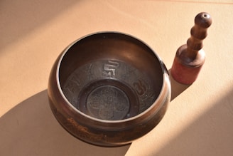 A metal singing bowl with intricate designs rests on a surface, accompanied by a wooden mallet with a rounded handle. The setup is illuminated by natural light, creating soft shadows.