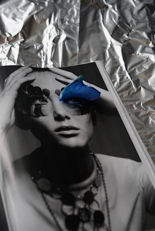 A monochrome photograph of a person whose eyes are obscured by abstract shapes, possibly pieces of nature or art. A vibrant blue petal is placed on the page, adding a striking contrast. The background appears to be crumpled aluminum foil, creating a textured, reflective backdrop.