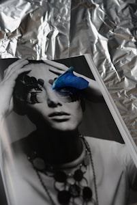 A monochrome photograph of a person whose eyes are obscured by abstract shapes, possibly pieces of nature or art. A vibrant blue petal is placed on the page, adding a striking contrast. The background appears to be crumpled aluminum foil, creating a textured, reflective backdrop.