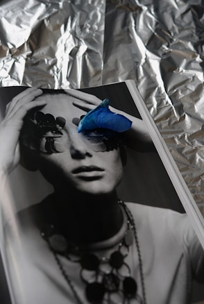 A monochrome photograph of a person whose eyes are obscured by abstract shapes, possibly pieces of nature or art. A vibrant blue petal is placed on the page, adding a striking contrast. The background appears to be crumpled aluminum foil, creating a textured, reflective backdrop.