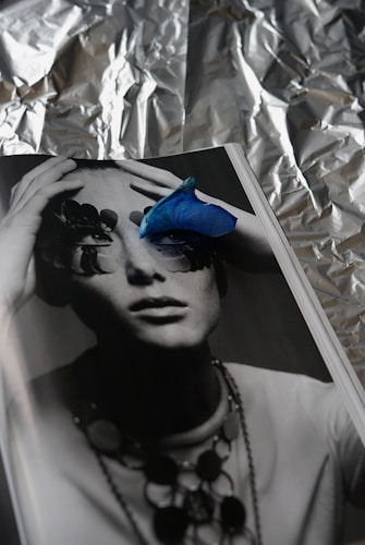 A monochrome photograph of a person whose eyes are obscured by abstract shapes, possibly pieces of nature or art. A vibrant blue petal is placed on the page, adding a striking contrast. The background appears to be crumpled aluminum foil, creating a textured, reflective backdrop.