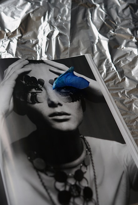 A monochrome photograph of a person whose eyes are obscured by abstract shapes, possibly pieces of nature or art. A vibrant blue petal is placed on the page, adding a striking contrast. The background appears to be crumpled aluminum foil, creating a textured, reflective backdrop.