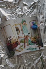 A fashion magazine is open to a page featuring a model in a stylish, patterned turquoise outfit and matching hat. The setting is an ornate and luxurious room with gold-accented furniture, a gilded mirror, and floral decorations. A multicolored rose lies on the pages. The background features crumpled reflective silver foil.