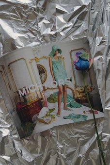 A fashion magazine is open to a page featuring a model in a stylish, patterned turquoise outfit and matching hat. The setting is an ornate and luxurious room with gold-accented furniture, a gilded mirror, and floral decorations. A multicolored rose lies on the pages. The background features crumpled reflective silver foil.