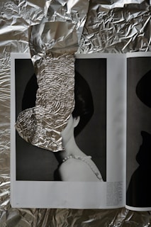 An open magazine rests on a crumpled sheet of aluminum foil. The magazine displays a monochrome photograph of a woman wearing a pearl necklace, partially obscured by a piece of crumpled foil strategically placed over her face. The juxtaposition of the shiny foil against the black and white photo creates an intriguing visual contrast.