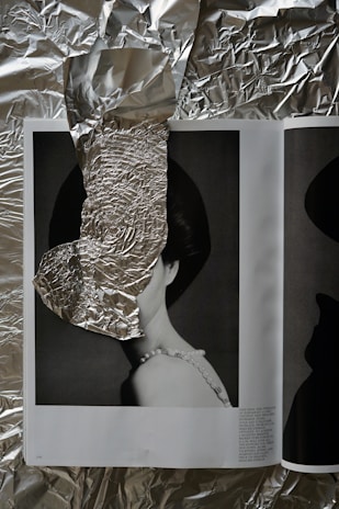 An open magazine rests on a crumpled sheet of aluminum foil. The magazine displays a monochrome photograph of a woman wearing a pearl necklace, partially obscured by a piece of crumpled foil strategically placed over her face. The juxtaposition of the shiny foil against the black and white photo creates an intriguing visual contrast.