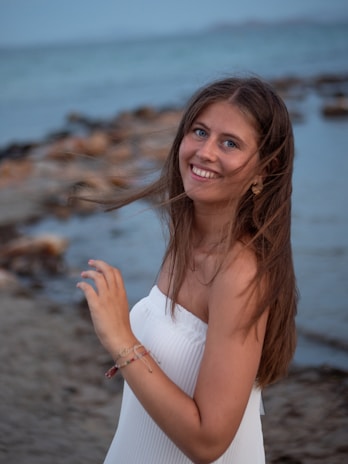 Smiling woman wearing a shell bracelet, standing by a rocky coastline under a bright blue sky.