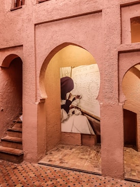 A textured, terracotta-colored architectural interior featuring arches and a small stairway. A photograph of a person engaged in intricate artistic work is displayed on one of the walls.