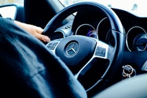 A close-up view of a person's hand gripping the steering wheel of a Mercedes-Benz car. The image focuses on the wheel's Mercedes logo and dashboard instruments, providing a sense of driving and control. The key ring with keys is visible in the ignition.