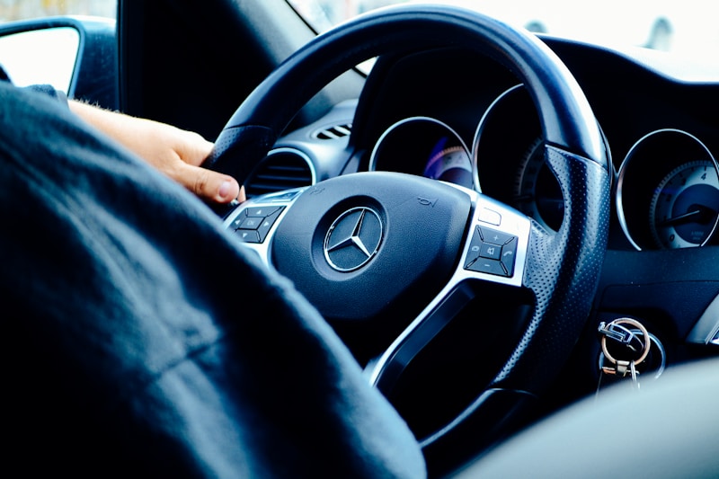A close-up view of a person's hand gripping the steering wheel of a Mercedes-Benz car. The image focuses on the wheel's Mercedes logo and dashboard instruments, providing a sense of driving and control. The key ring with keys is visible in the ignition.