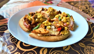 Three small pizzas on a white plate, each topped with grilled chicken, corn, and vegetables such as bell peppers and zucchini. The plate is set on a decorative tablecloth with intricate swirling patterns in brown and gold tones.