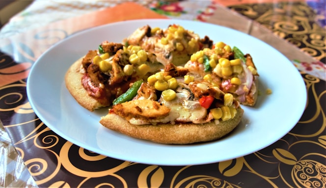 Three small pizzas on a white plate, each topped with grilled chicken, corn, and vegetables such as bell peppers and zucchini. The plate is set on a decorative tablecloth with intricate swirling patterns in brown and gold tones.