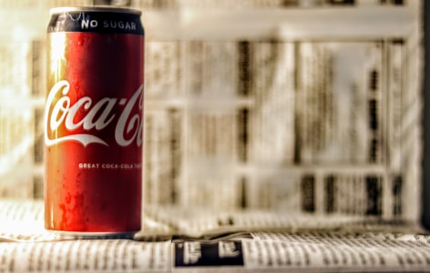 A Coca-Cola can with 'No Sugar' printed at the top is placed against a background of blurred text. The can is prominently featured in vibrant red color with white logo and details.