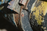 A close-up view of a weathered and corroded metal surface showing peeling paint and rust. The surface features bolts and layers of aged material with a rough texture.