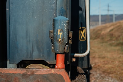 A close-up of an industrial object with a prominent blue casing featuring a yellow lightning bolt symbol. A red cylindrical part is visible below the blue component. The background includes a metallic surface and a distant view of a grassy area with power lines.